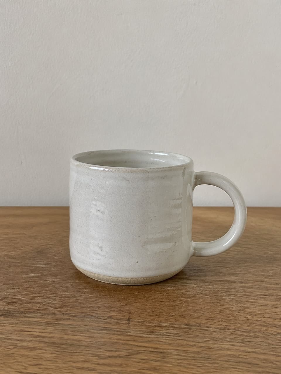 Real handmade cream ceramic mug on a wooden table, with throwing ridges, iron specks, and an unglazed clay foot